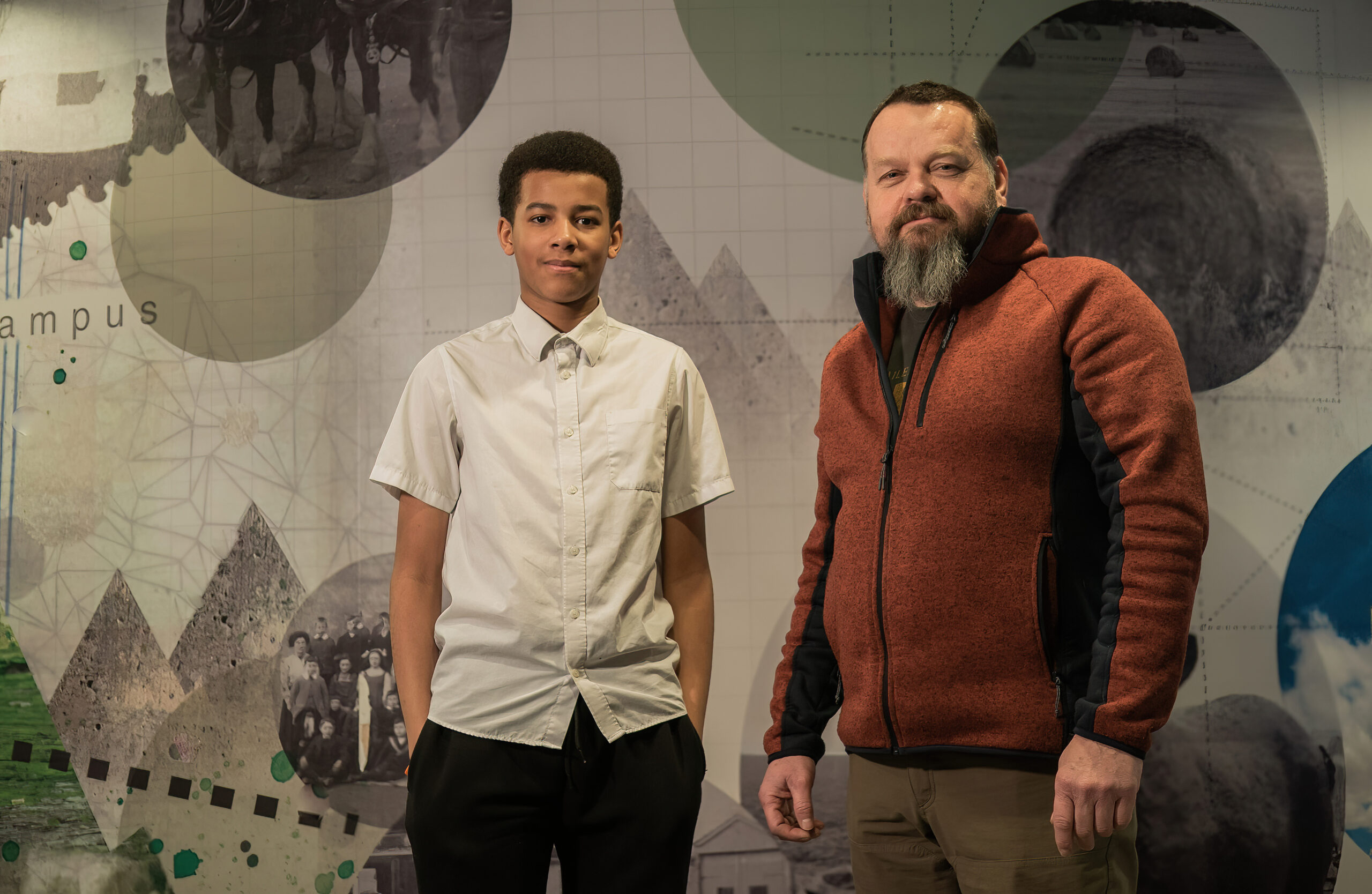 picture of a male mentor and his male mentee posing behind a patterned wallpaper in a school.