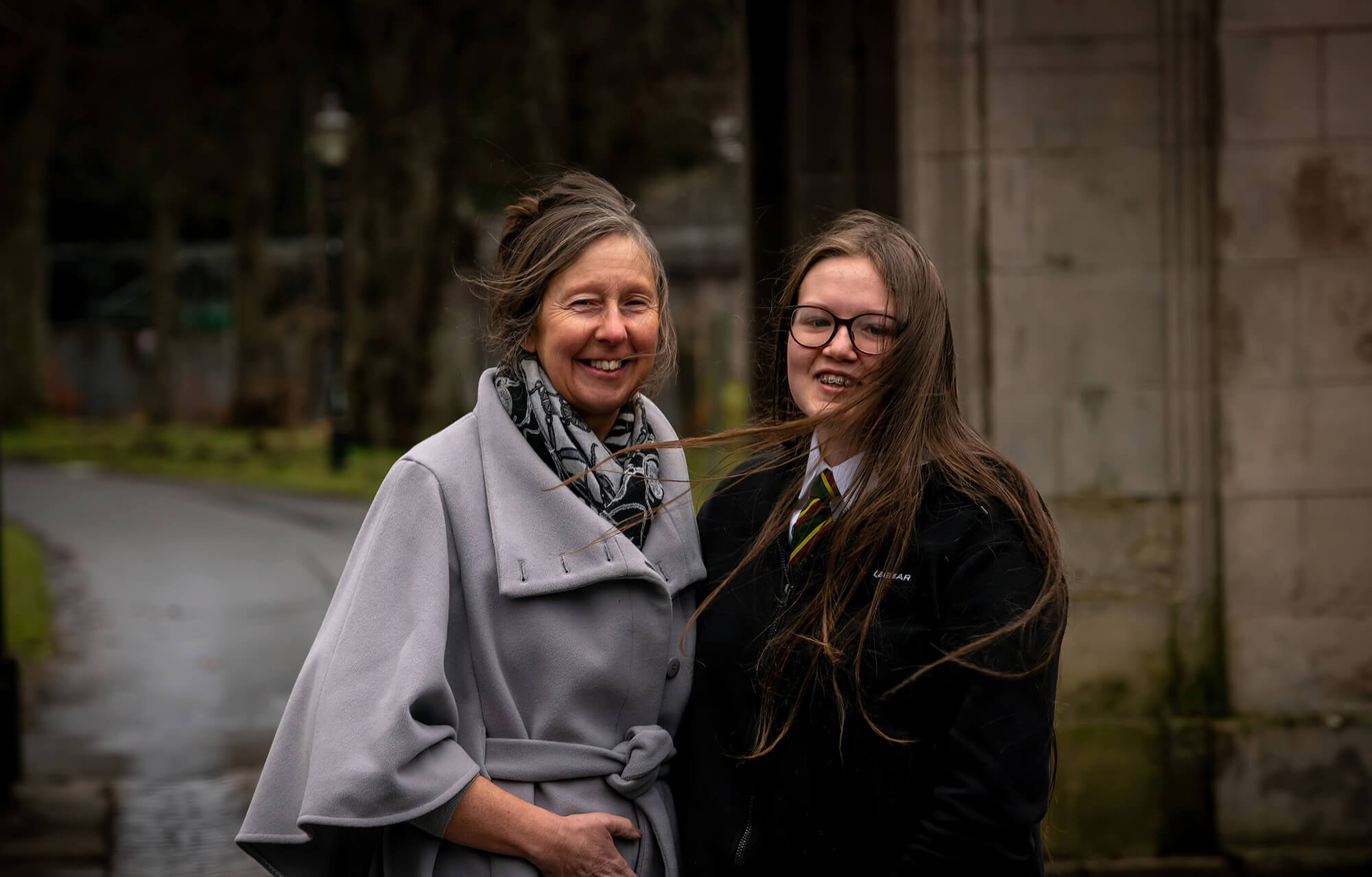 Two people standing outdoors on a windy day, smiling at the camera. One wears a light grey coat and scarf, the other a school uniform with glasses and long hair blowing in the wind.