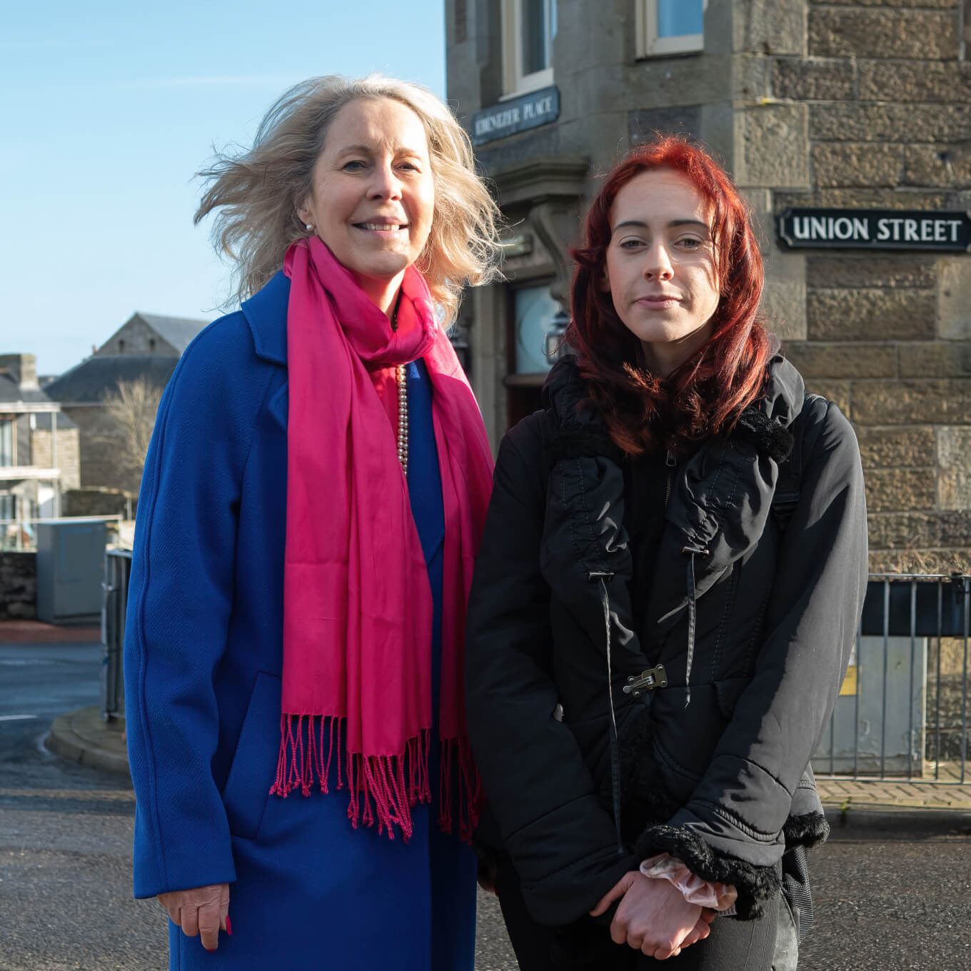 Two people stand outside wearing outdoor clothes.