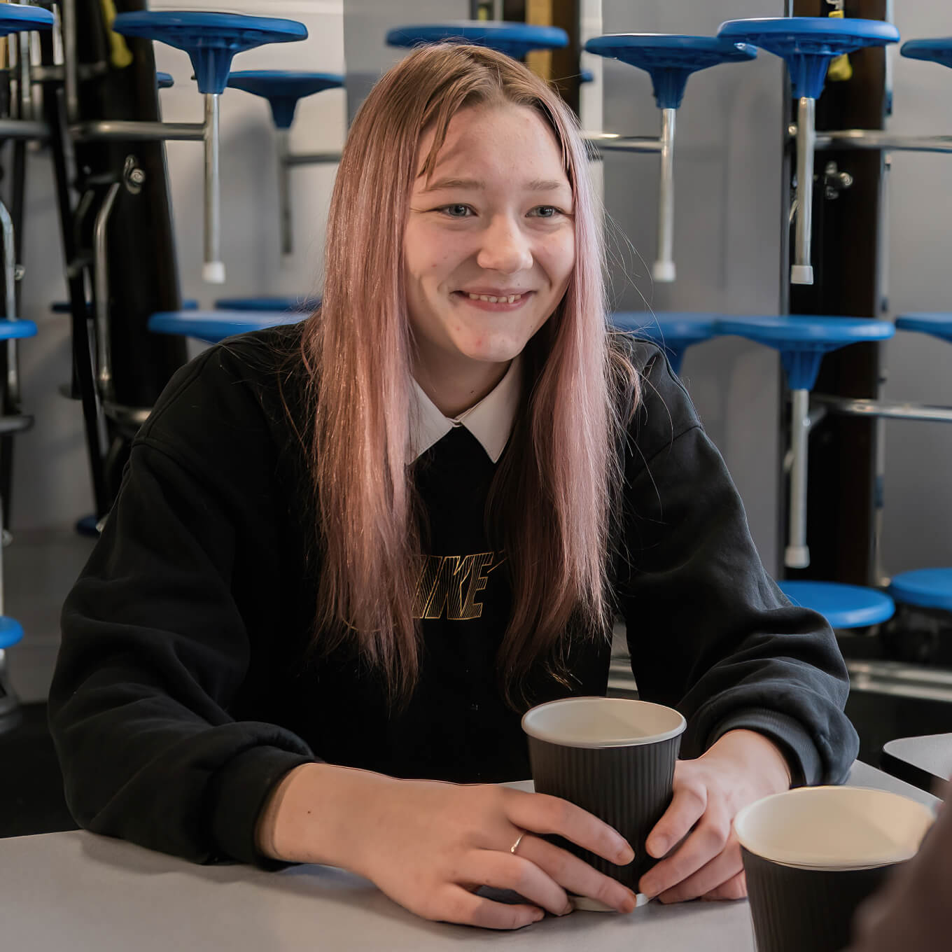 A young person with long pink hair sits at a table holding a paper cup, smiling in a room with stacked blue stools in the background.